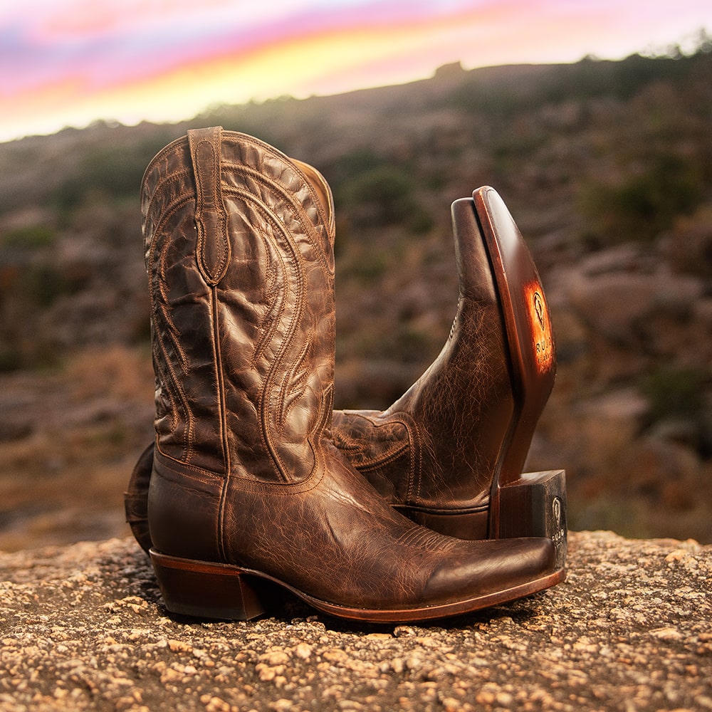 A pair of RUJO Mad Dog Leather cowboy boots resting on a rock at sunset