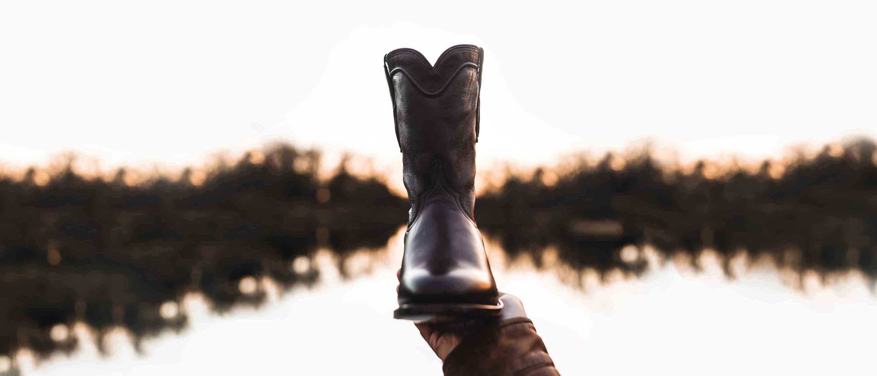 Man holding a RUJO Caravan Calfskin cowboy boot up for inspection by a lake at sunset