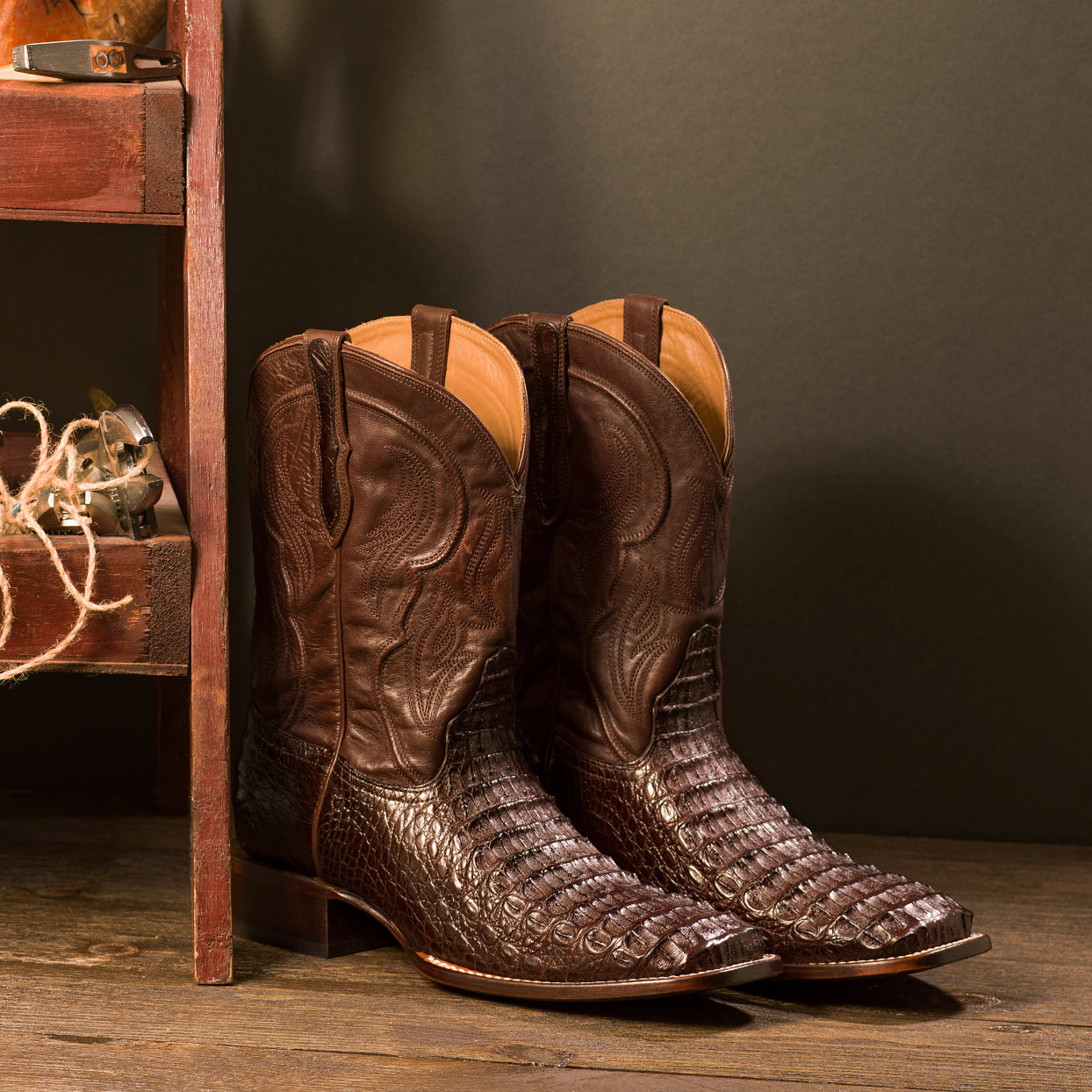 A pair of RUJO caiman cowboy boots next to a wooden shelf